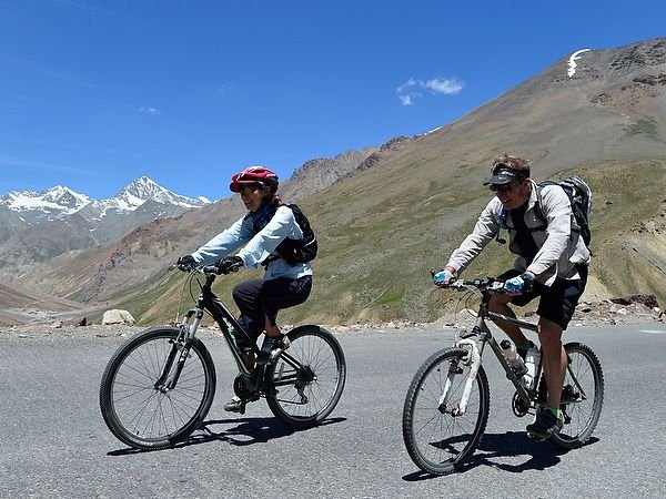Two cyclists ride mountain bikes on a scenic road with mountains in the background.