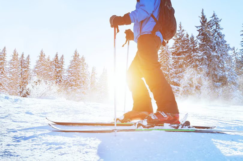 Skier in blue jacket on snowy slope with sun shining through pine trees