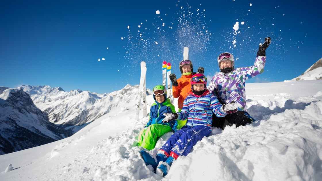 Group of skiers in colorful gear on a snowy mountain, smiling and capturing memories under a clear blue sky.