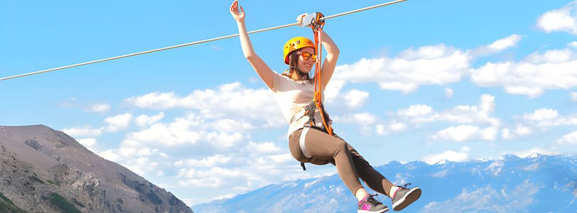 Woman ziplining high above mountain peaks under a clear blue sky, enjoying an adventurous outdoor experience.